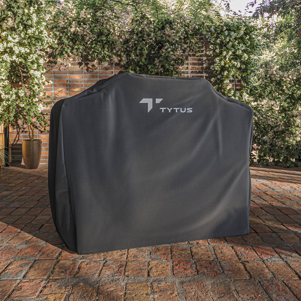 TYTUS island grill cover, protecting a grill on a brick patio with flowering bushes in the background.